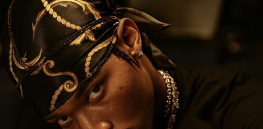 黒いバンダナとジュエリーを身につけたラッパーが床に座り、真剣な眼差しを向けるヒップホップアーティストのポートレート・d4vd