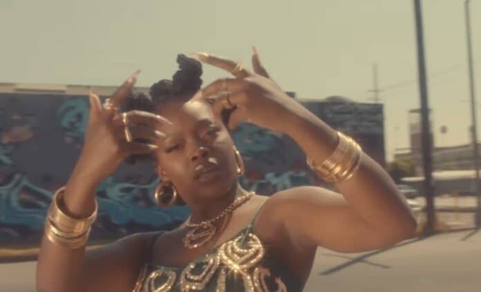 Charm La’Donnaが『Queen』のミュージックビデオで、ロサンゼルスのストリートを背景にハンドサインを掲げる瞬間。金のアクセサリーとエメラルドグリーンのドレスが太陽光に輝き、ヒップホップとアフリカン・フェミニズムの融合を象徴している。