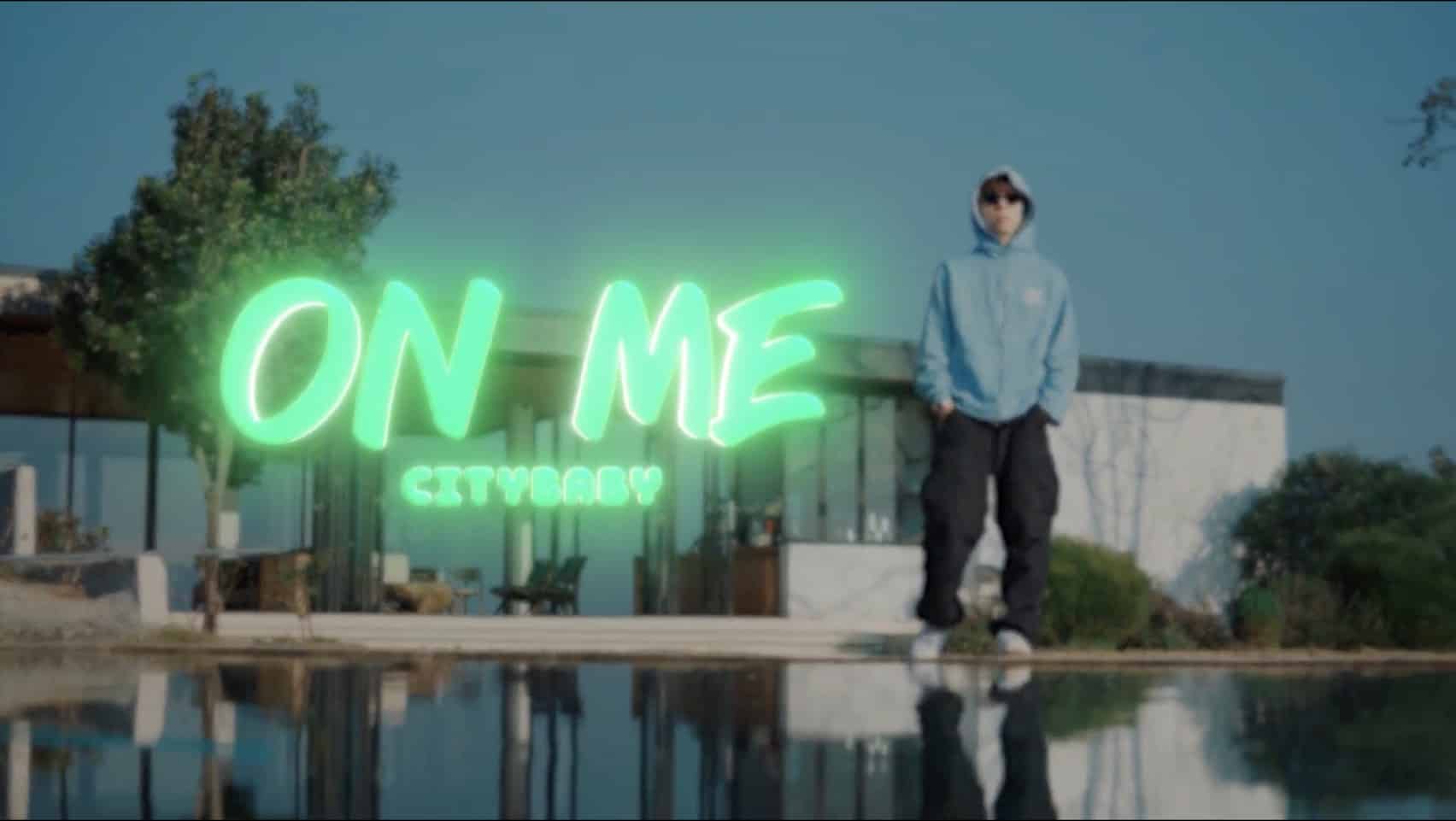 「On Me」MVでネオングリーンの「ON ME」サインの前、水辺のガラス張りの家を背にライトブルーのフーディーで立つCityBaby