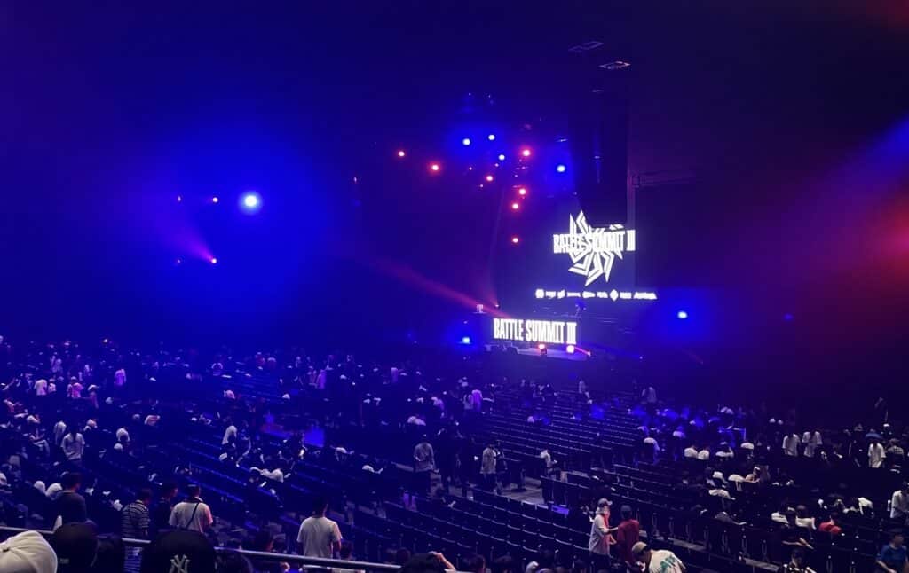 2026年8月11日のBATTLE SUMMIT III、Kアリーナ横浜の客席から見たステージ。大型スクリーンとステージ前面に大会ロゴが表示され、青と赤の照明が客席まで届いている
