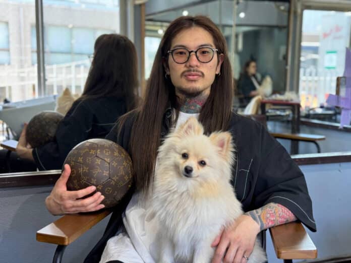 サロンの椅子に座り、白い犬を膝に抱くヘアアーティストのBraidKingNori
