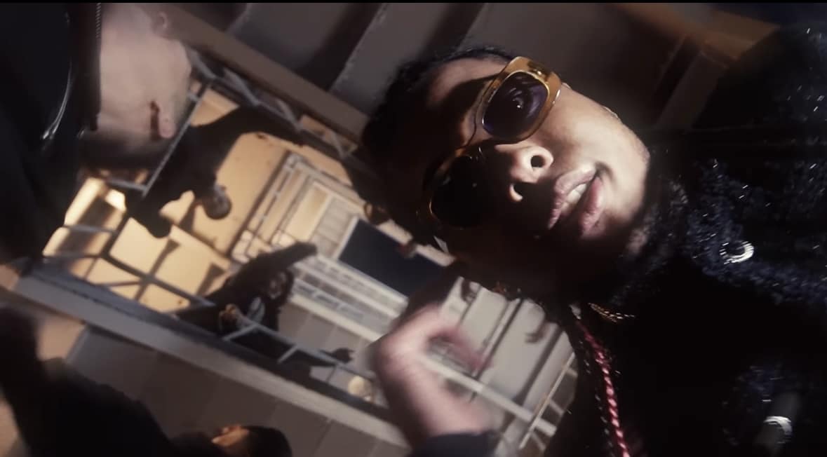 BLAISE wearing sunglasses in a tilted close-up with buildings behind him