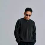 黒いニットとサングラスを着用し、グレーの背景の前に立つDJ KANJI（取材先提供）