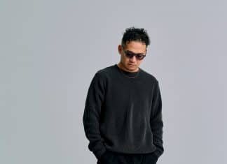 黒いニットとサングラスを着用し、グレーの背景の前に立つDJ KANJI（取材先提供）