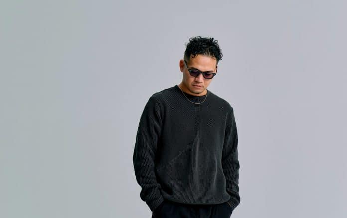 黒いニットとサングラスを着用し、グレーの背景の前に立つDJ KANJI（取材先提供）
