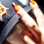 Close-up of SEDY NEZZ in a dark cap, holding two fingers with flame-pattern nails near the camera