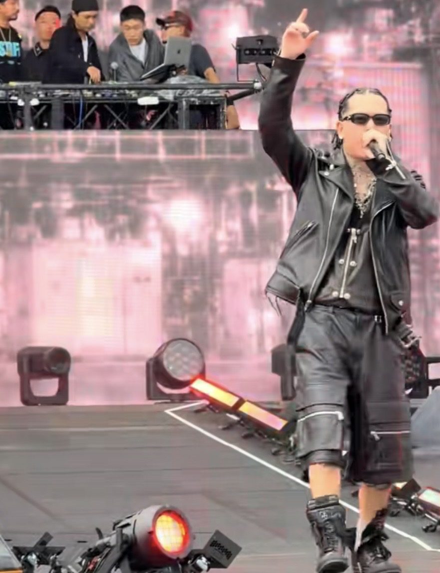 黒いレザーのジャケットとショーツ姿で、片手を上げてラップするYZERR。背後に一段高いDJブースが見える。