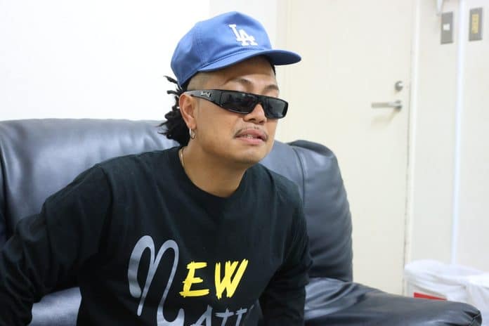 青いLAキャップと黒いサングラスを着用し、ソファに座るMAIKI BOOGIE