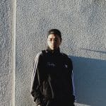 REDDY standing by a sunlit wall in a black track jacket, glasses and a backward cap