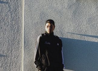 黒いトラックジャケット、眼鏡、後ろ向きのキャップ姿で、日差しの当たる壁の前に立つREDDY