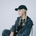 黒いキャップに金髪の三つ編み、ネイビーのセットアップでスツールに座るラッパーのyummma