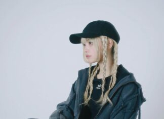 黒いキャップに金髪の三つ編み、ネイビーのセットアップでスツールに座るラッパーのyummma